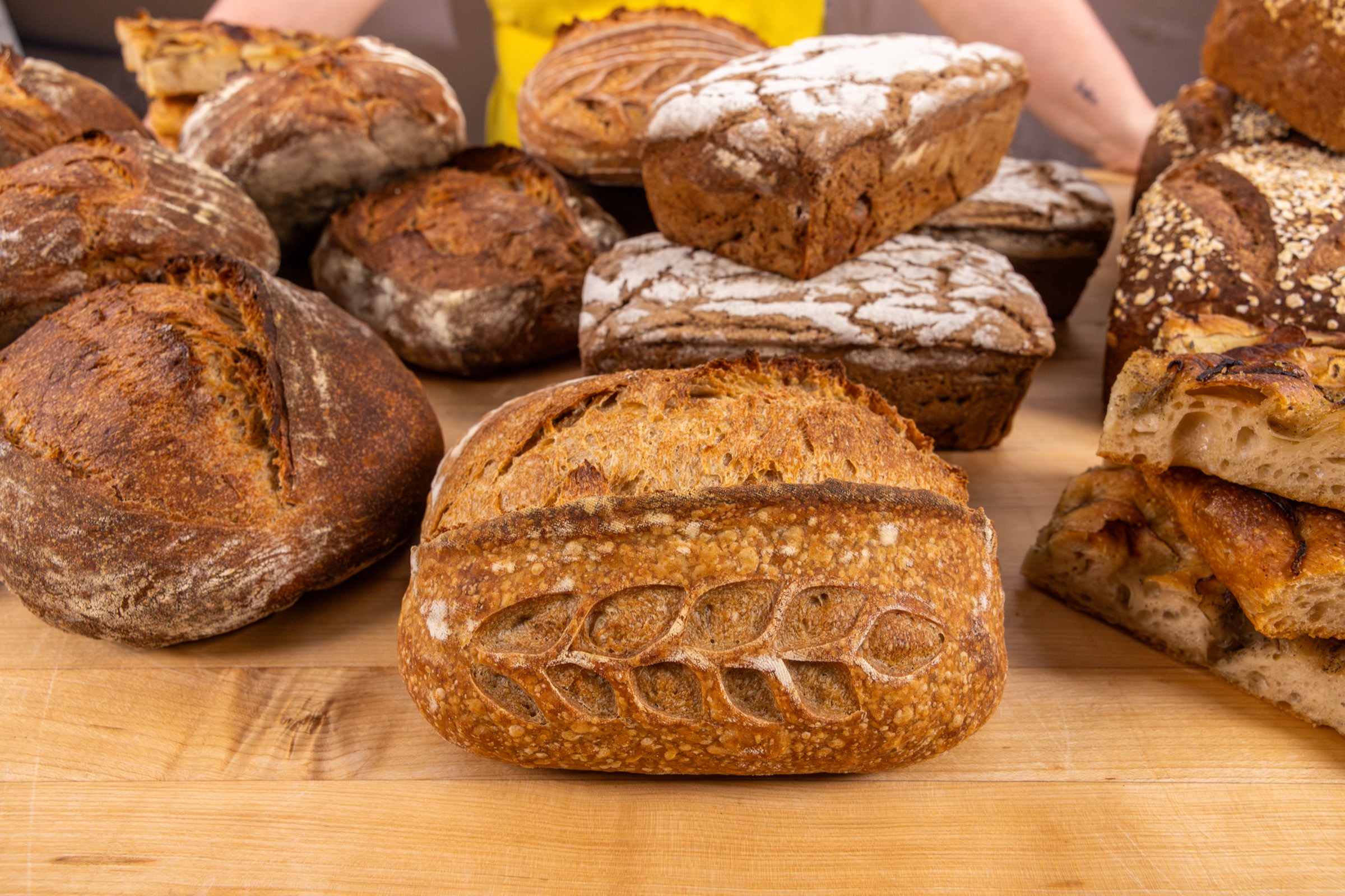 A wooden table crowded with fresh sourdough — scored country loaves, dusted rye pan loaves, oat-topped batards, and slabs of focaccia