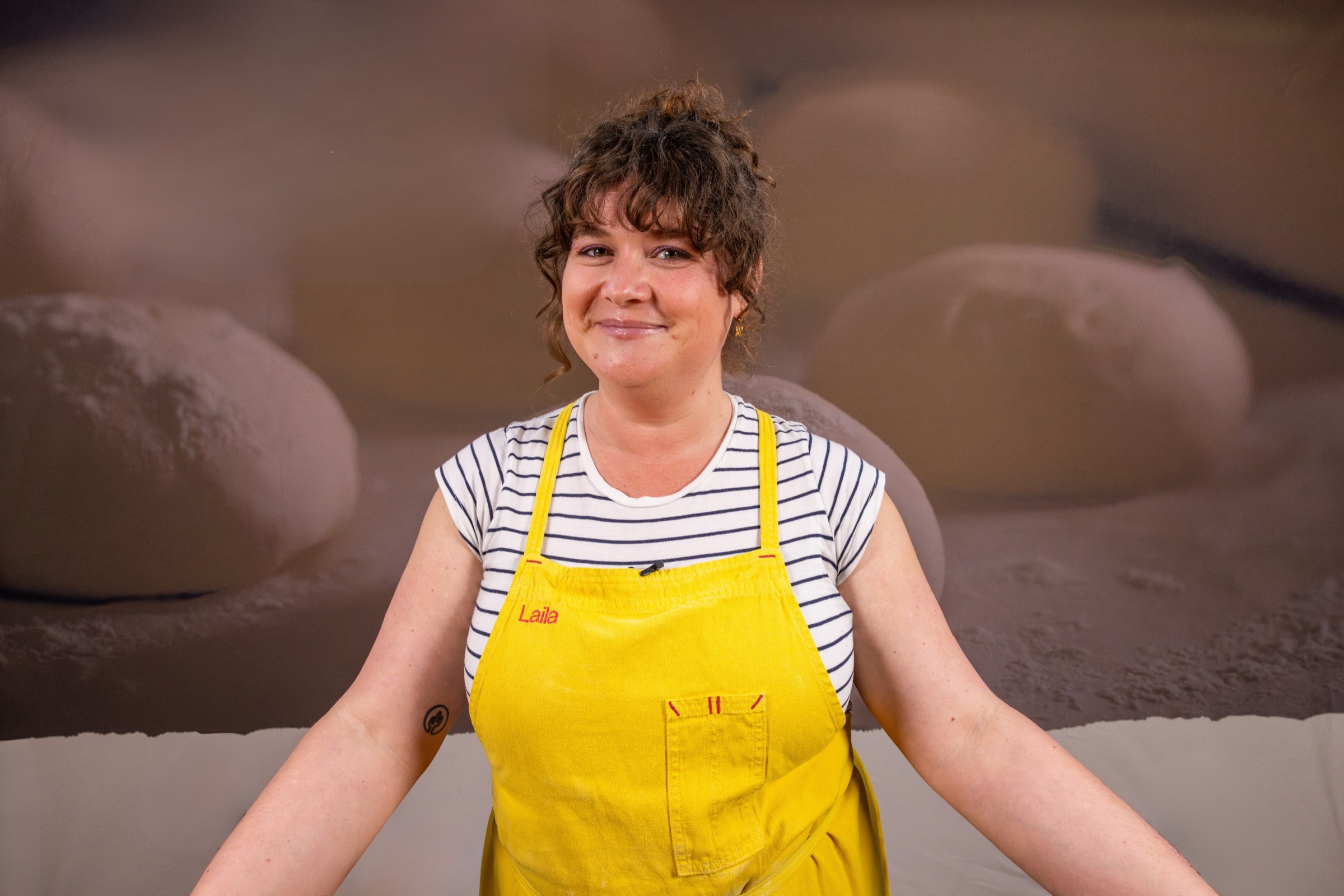Laila in her yellow baker's apron embroidered with her name, smiling in front of rounds of proofing dough