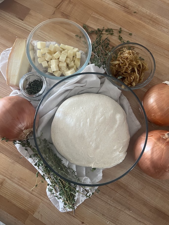Everything you need for french onion sourdough laid out on a butcher block: a bowl of dough, cubed Gruyère, a jar of chopped thyme, caramelized onions, fresh thyme sprigs, and whole sweet onions