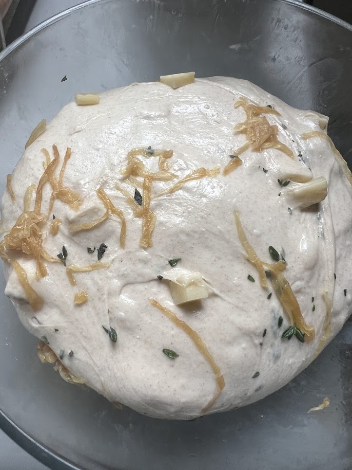 Dough in a glass bowl topped with strands of caramelized onion, cubes of Gruyère, and fresh thyme leaves, ready for the first stretch and fold